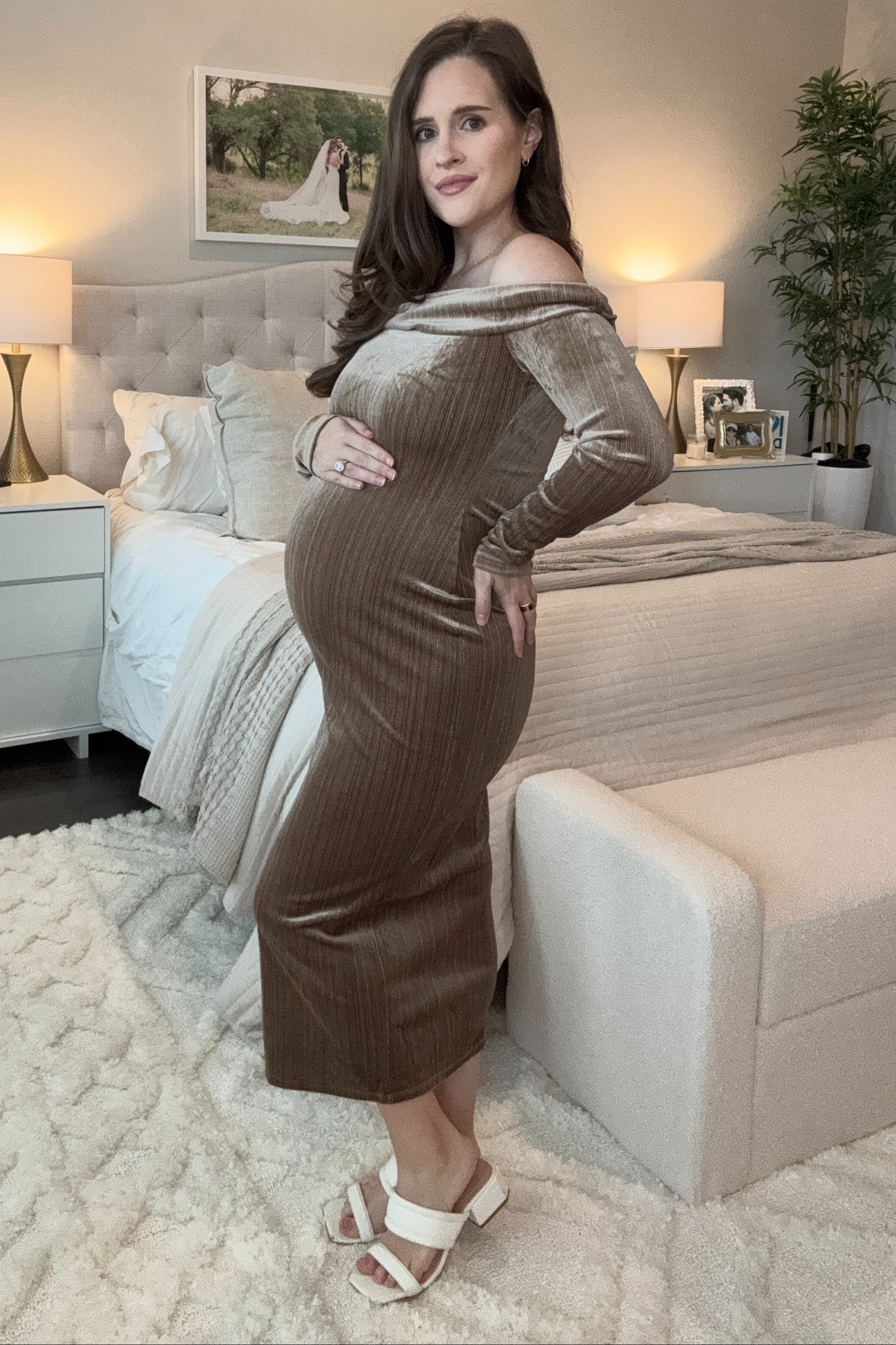 woman wearing a brown off-shoulder maternity dress with a visible baby bump, standing in a bedroom setting
