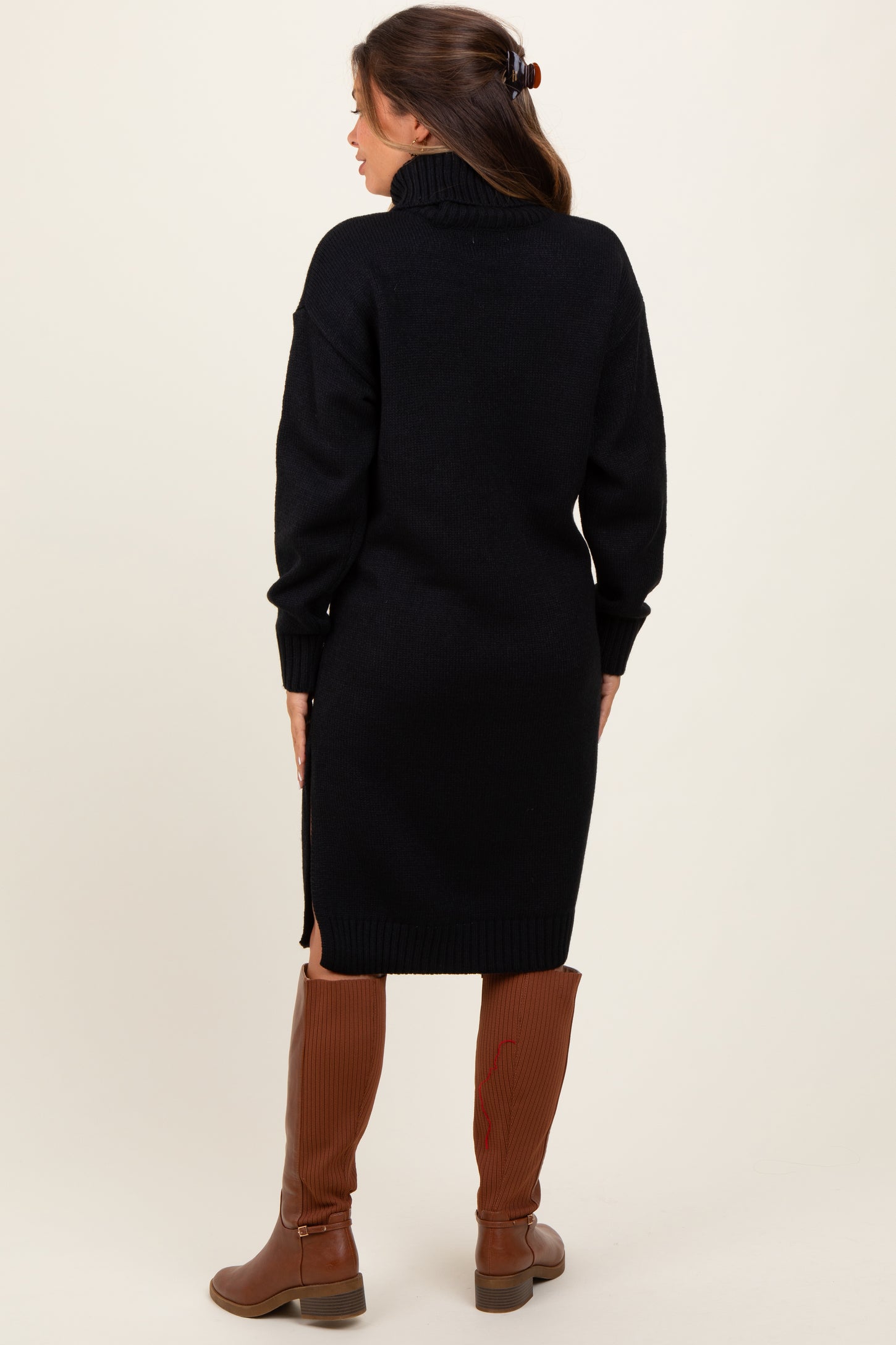 woman wearing a black maternity sweater dress with a high neckline and side slits, paired with brown knee-high boots