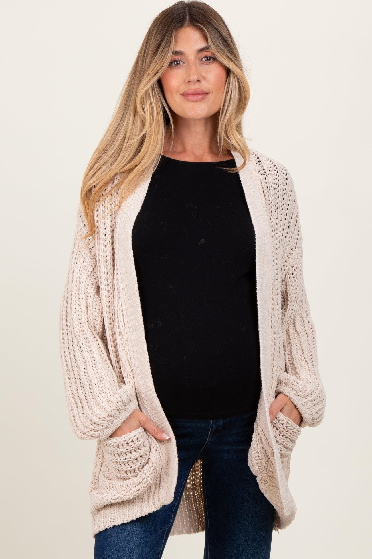 woman wearing a beige open-knit maternity cardigan with pockets and a black top, showing her baby bump