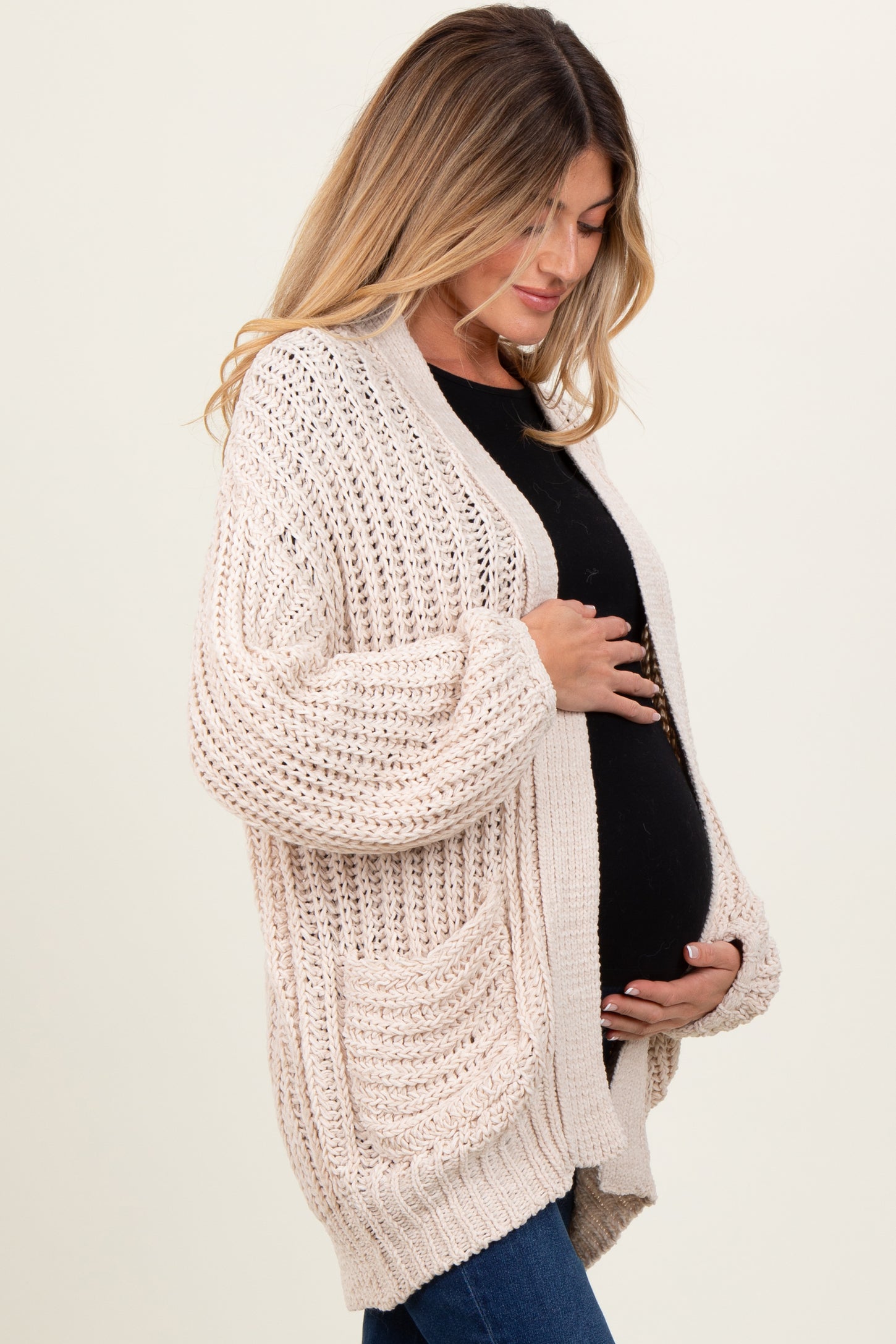 woman wearing a cream chunky knit maternity cardigan and gently holding her baby bump