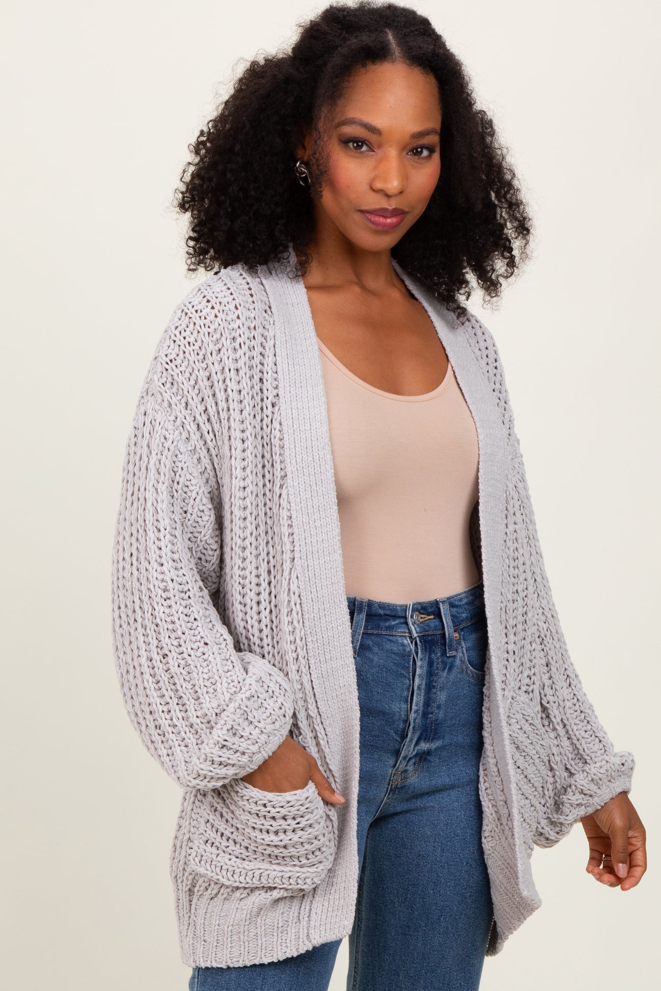woman wearing a light gray chunky knit cardigan with oversized sleeves and front pockets, paired with a beige top and jeans
