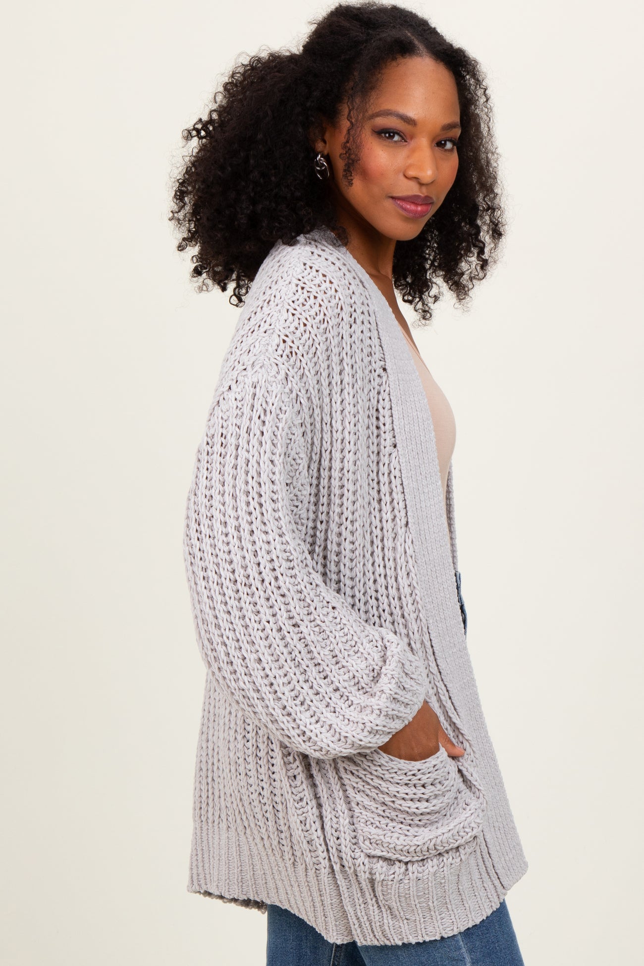 woman wearing a light gray chunky knit cardigan with pockets, paired with a beige top and blue jeans