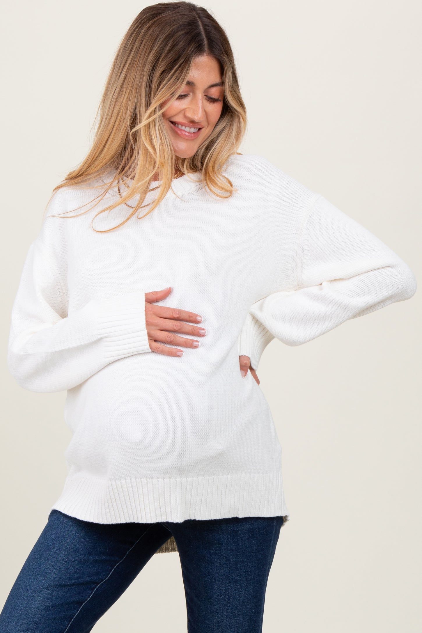 woman wearing a relaxed fit white maternity sweater and holding her baby bump, paired with denim jeans