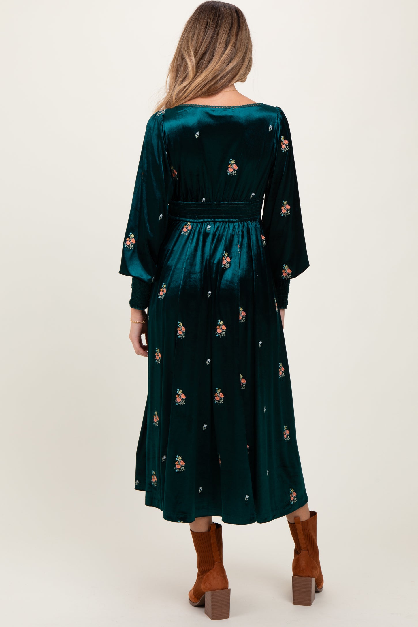 woman wearing a green velvet maternity dress with floral embroidery and long sleeves, standing with her back to the camera