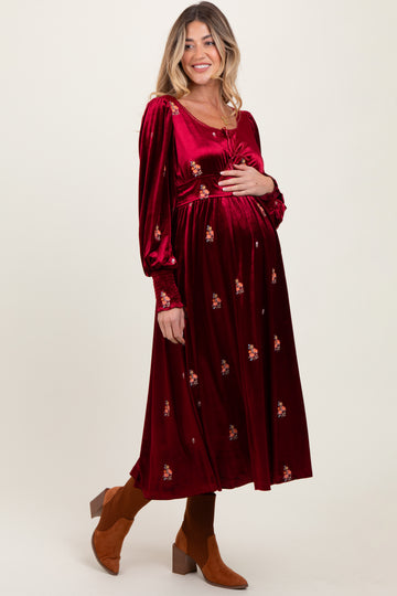 woman wearing a burgundy velvet maternity dress with floral embroidery and holding her baby bump