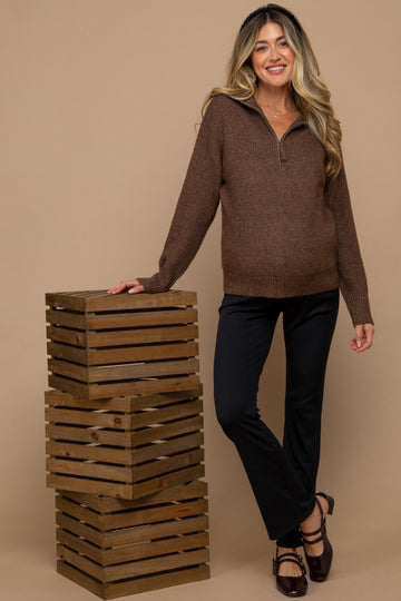 woman wearing a brown maternity sweater with a half zip and black pants, posing beside wooden crates with a visible baby bump