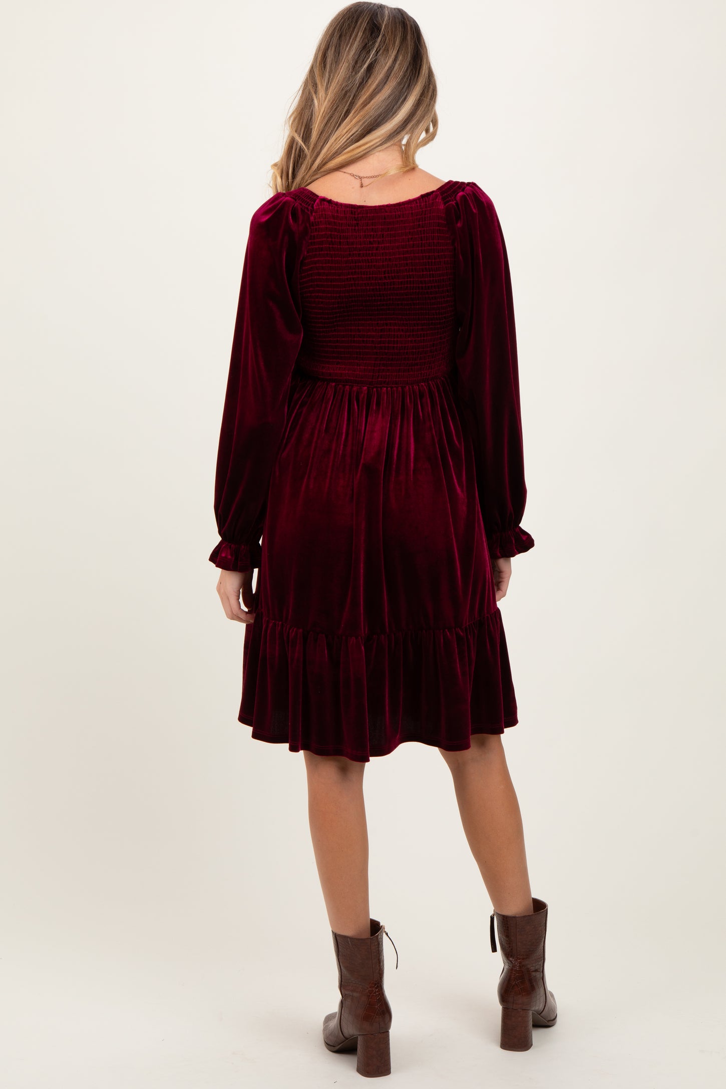 woman wearing a burgundy velvet maternity dress with long sleeves and a ruffled hem, showing a fitted smocked back