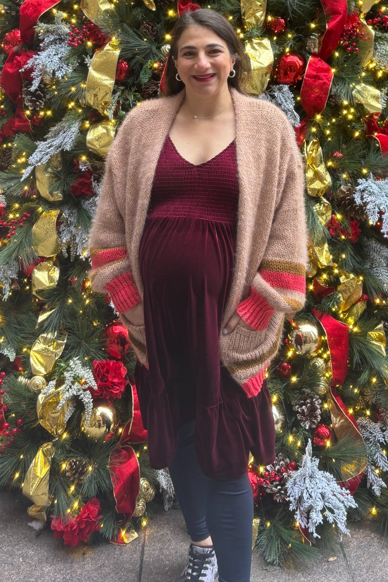 woman wearing a burgundy maternity dress and a beige oversized cardigan, posing with a baby bump in front of holiday decorations