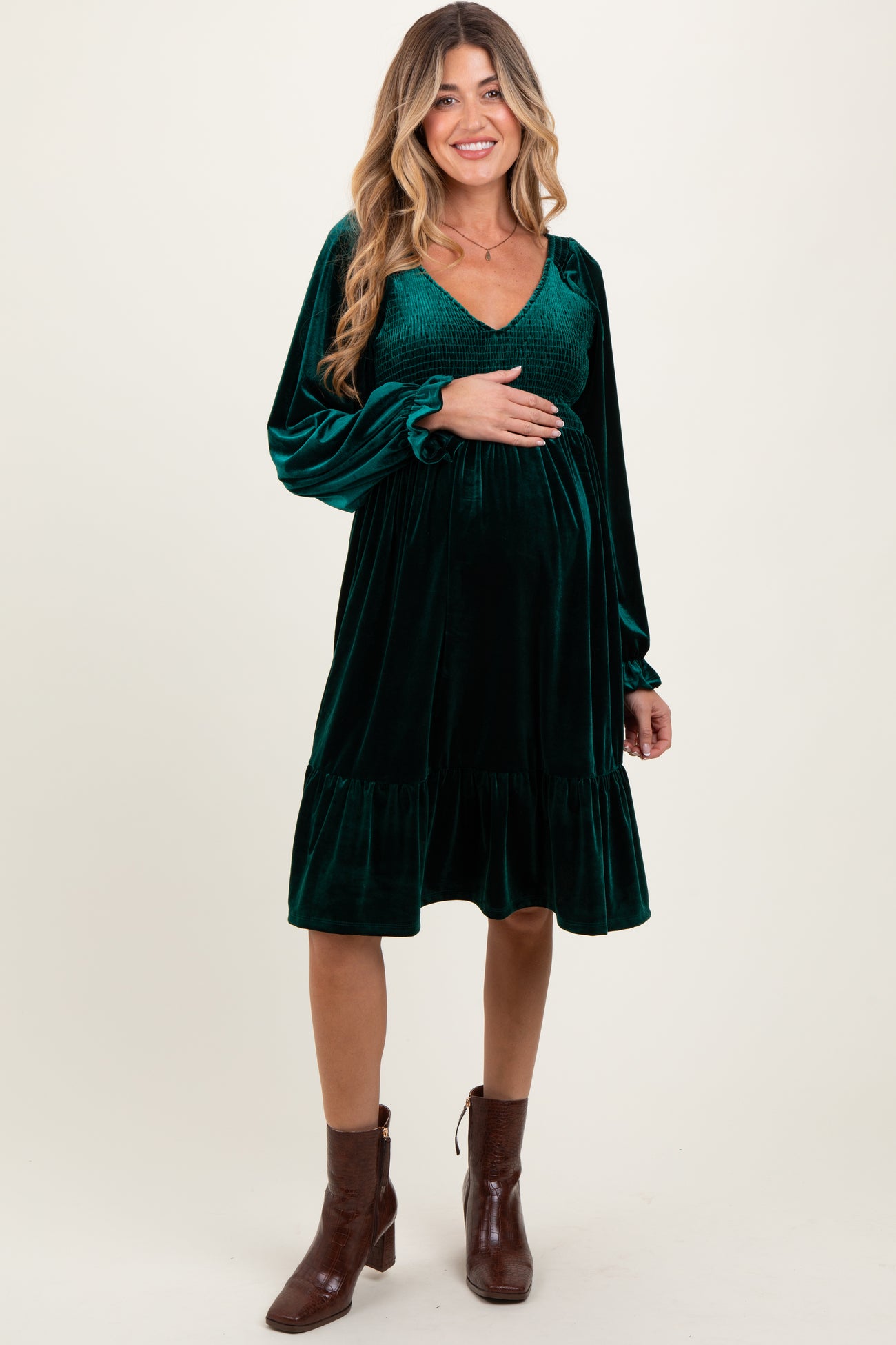 woman wearing a dark green velvet maternity dress with long sleeves and a ruffled hem, holding her baby bump