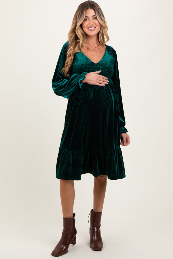 woman wearing a dark green velvet maternity dress with long sleeves and a ruffled hem, holding her baby bump
