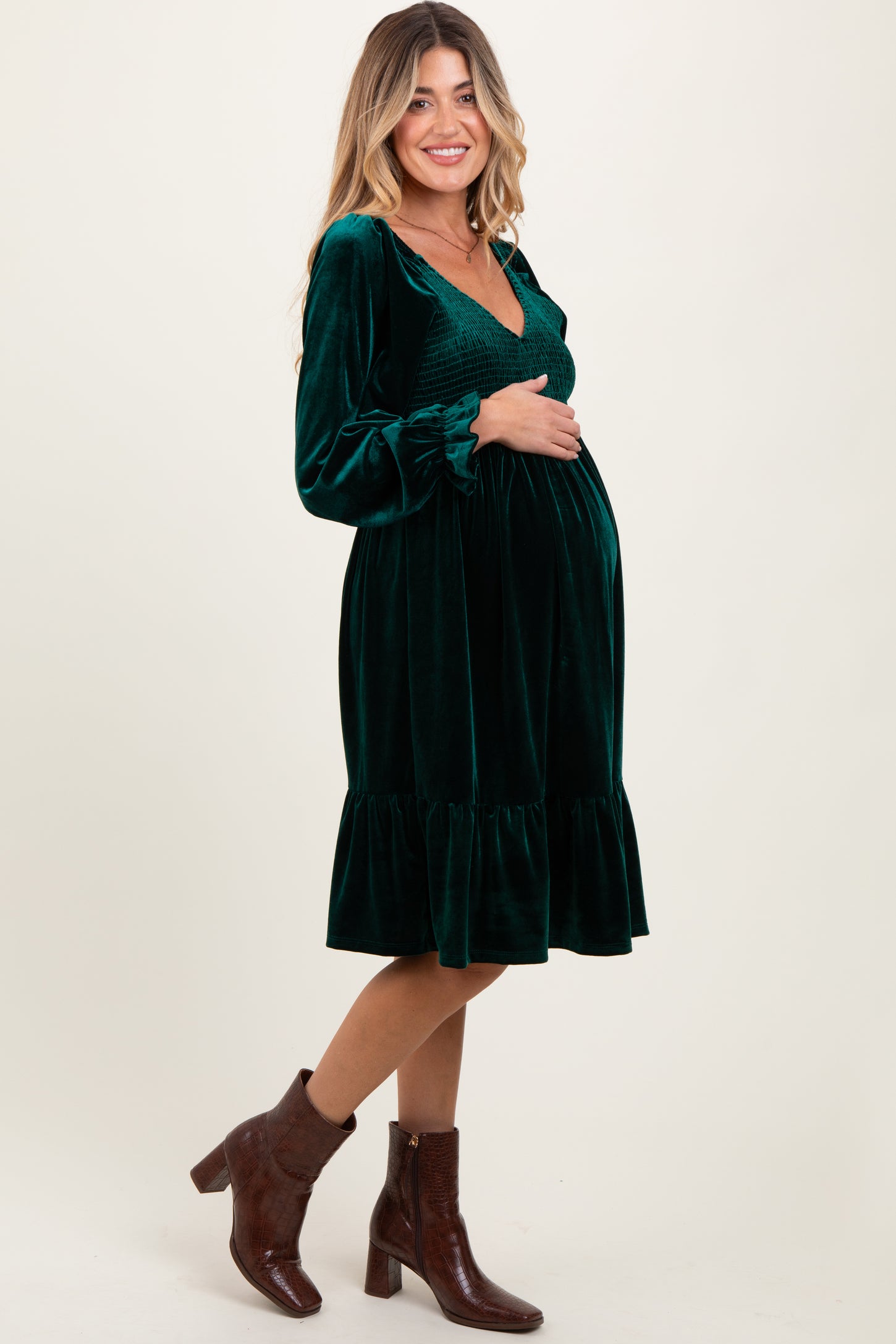 woman wearing a green velvet maternity dress with a smocked bodice and ruffle hem, holding her baby bump
