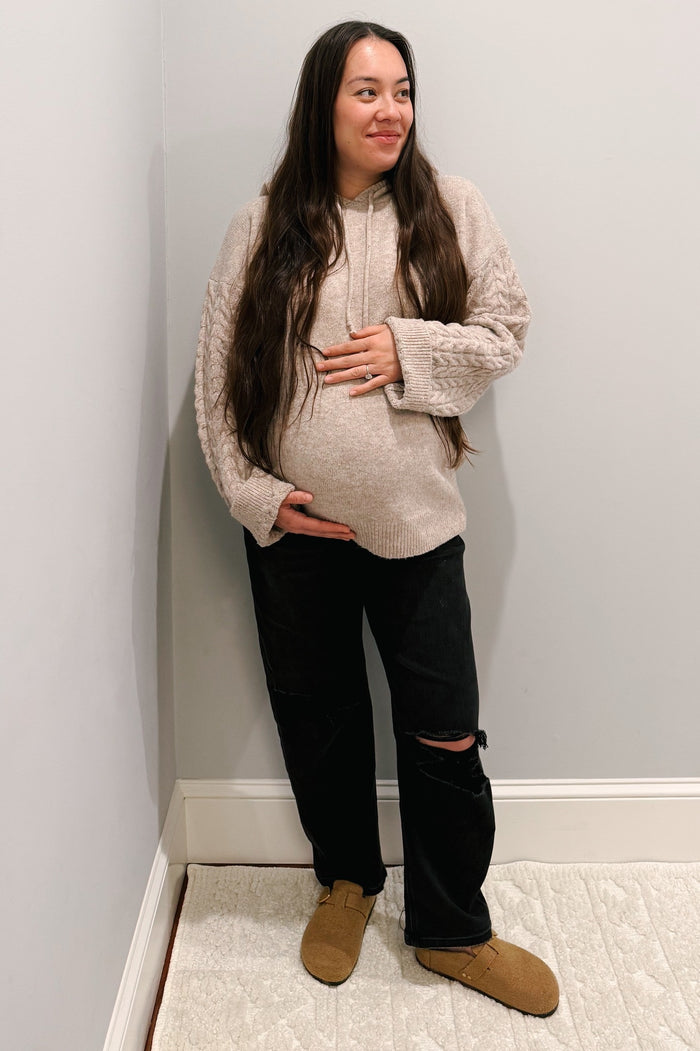 woman wearing a cream cable knit maternity hoodie and black ripped maternity jeans, holding her baby bump