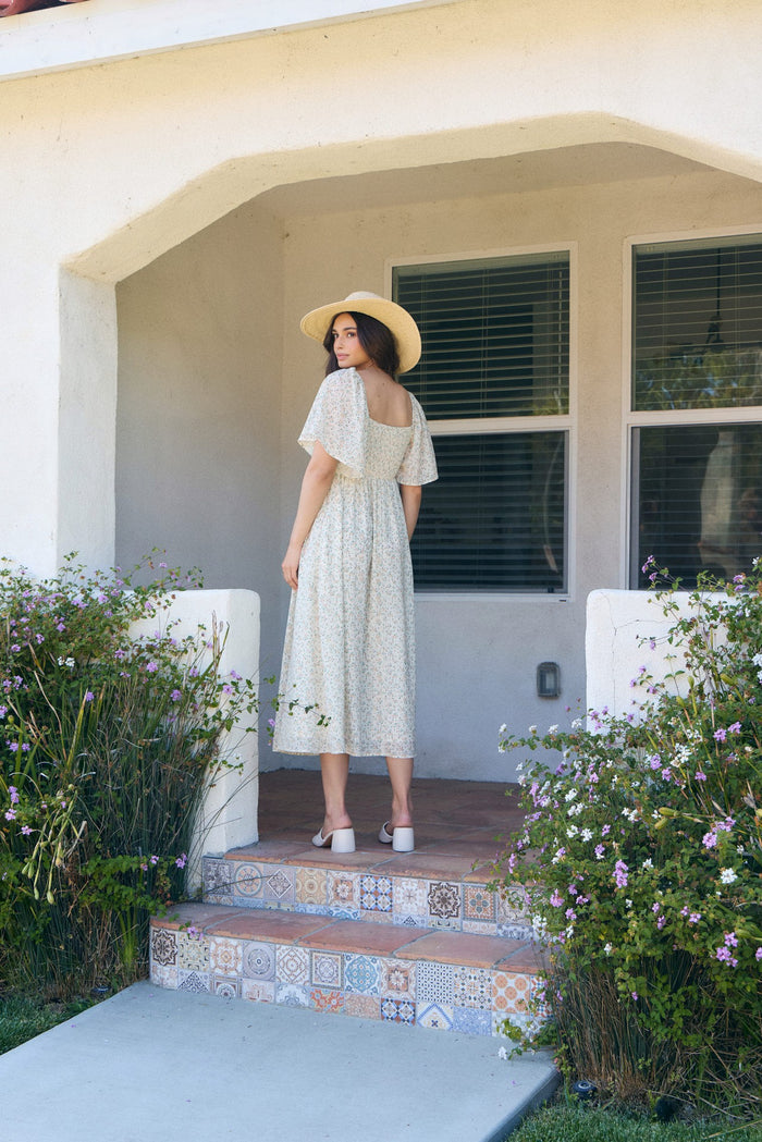 woman wearing a pale yellow floral midi dress with flutter sleeves and a wide-brimmed hat, standing on steps outside