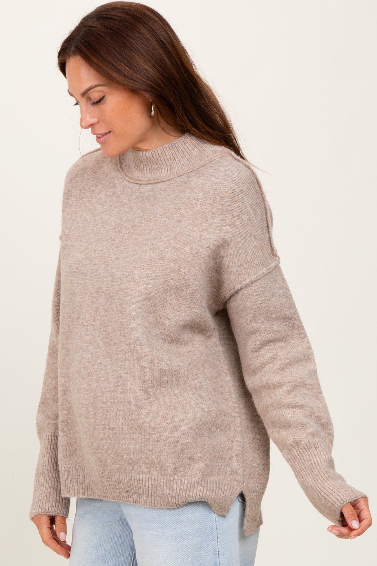 woman wearing a beige long sleeve sweater with a high neck and side slit detail, looking down and smiling