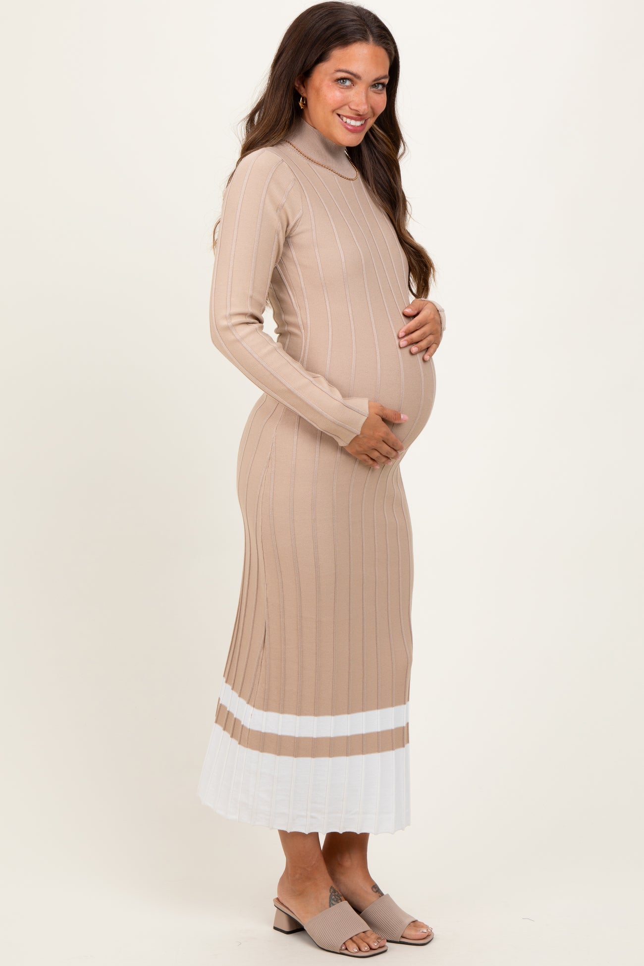woman wearing a beige ribbed maternity dress with white pleated hem and holding her baby bump