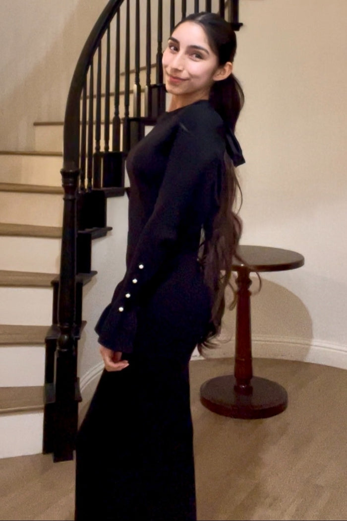 woman wearing a black long sleeve dress with a bow tie detail and button accents on the sleeves, standing by a staircase