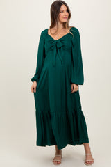 woman wearing a dark green maternity dress with a tie front and long sleeves, featuring a tiered skirt and a visible baby bump