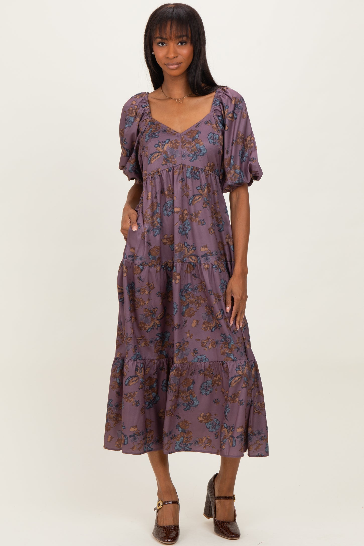 woman wearing a purple floral midi dress with puff sleeves and a tiered skirt, standing and smiling