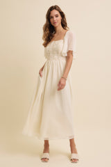 woman wearing an ivory maxi dress with floral detailing and flutter sleeves, standing against a neutral background