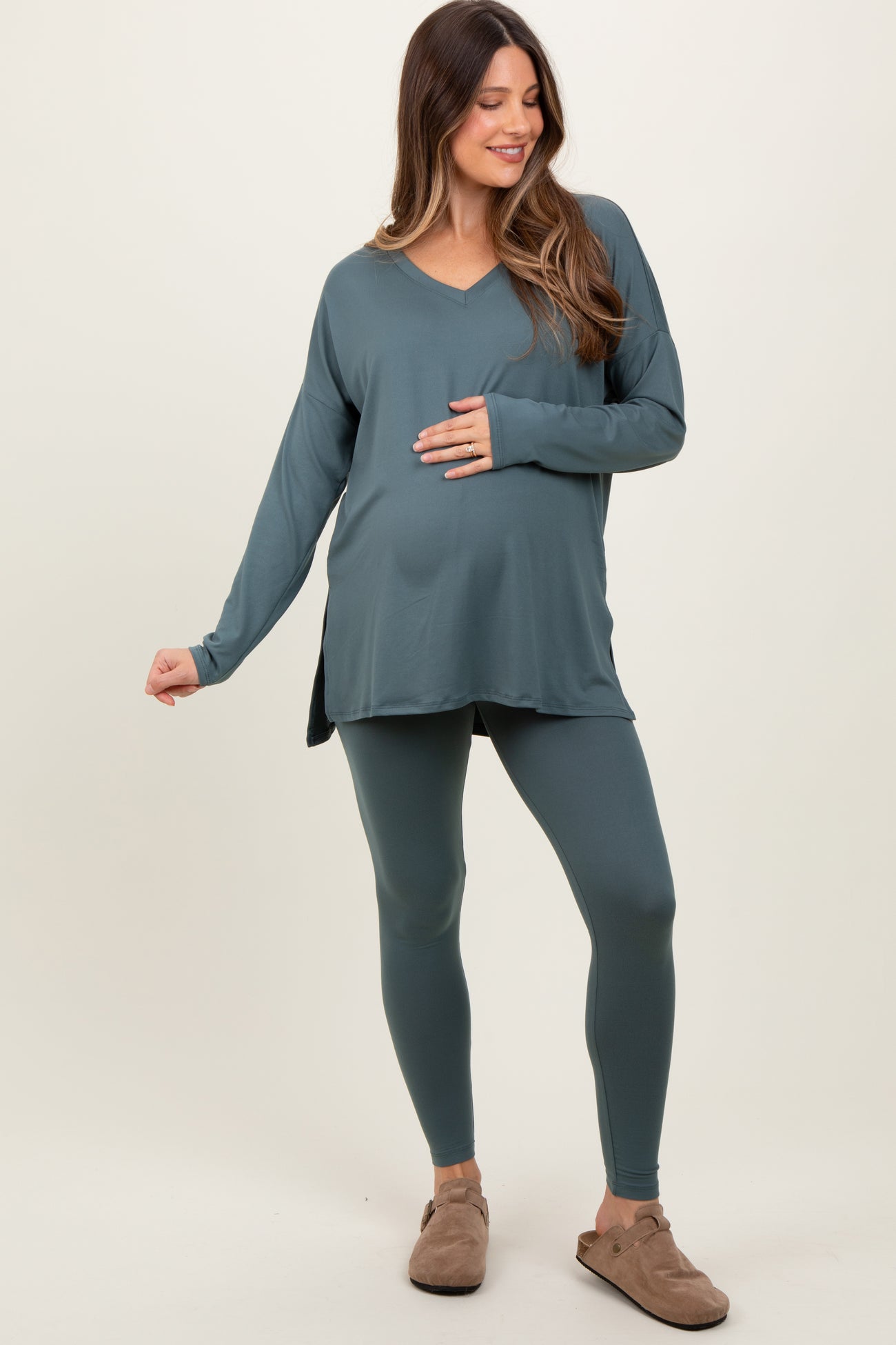 woman wearing a teal maternity top and matching leggings, gently holding her baby bump