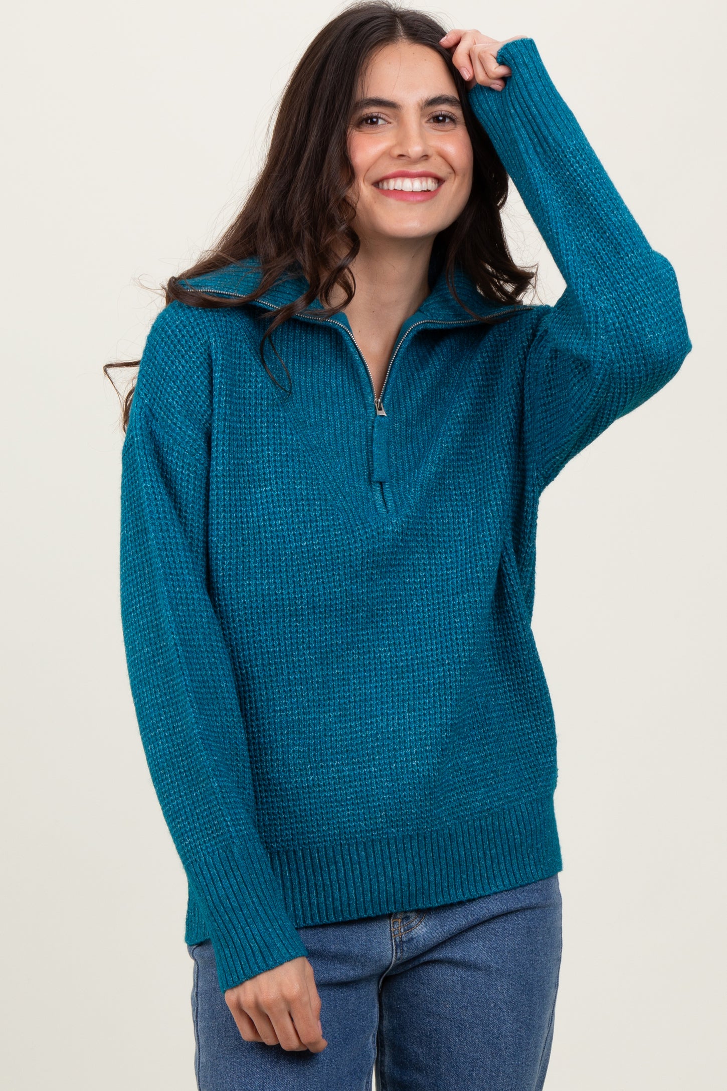 woman wearing a teal zip-up sweater with a textured knit design and a relaxed fit, smiling and posing with one hand in her hair