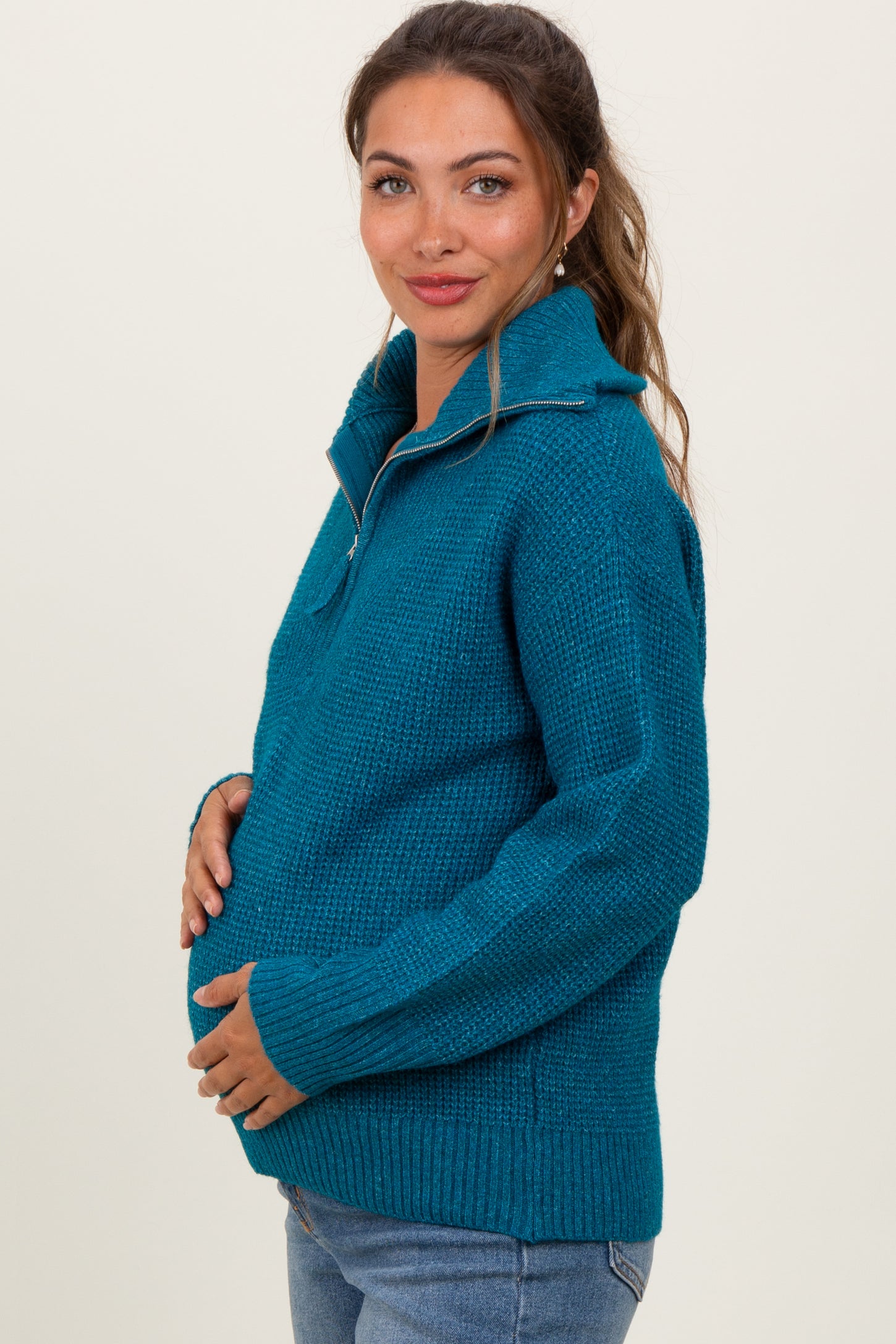 woman wearing a teal maternity sweater with a half zip and holding her baby bump