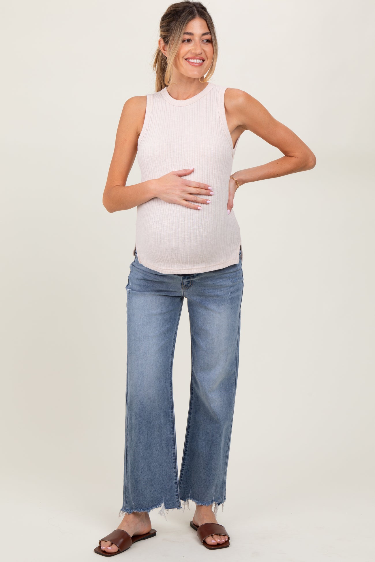 woman wearing a light pink ribbed maternity tank top and blue wide-leg jeans, holding her baby bump