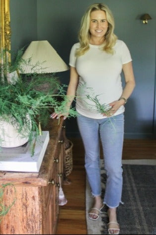 woman wearing a white short sleeve maternity top and blue jeans with a visible baby bump, posing indoors