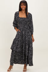 woman wearing a black floral maxi dress with long sleeves and a smocked bodice, standing with one hand on her hip