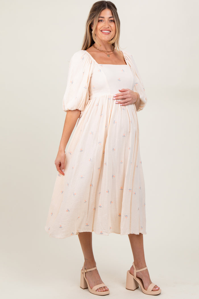 woman wearing a light pink maternity dress with puff sleeves and floral embroidery, holding her baby bump