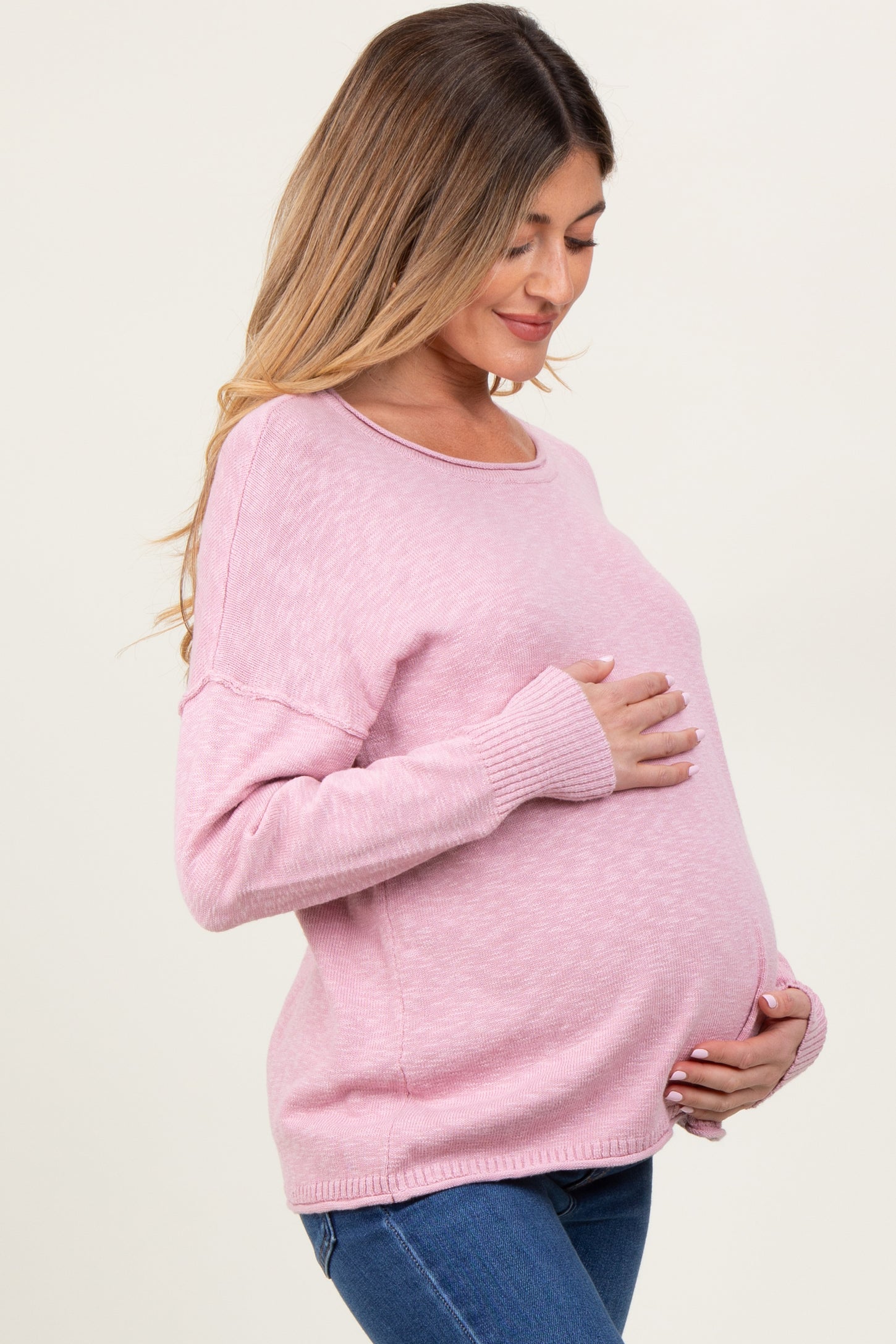 woman wearing a light pink maternity sweater, cradling her baby bump while posing sideways