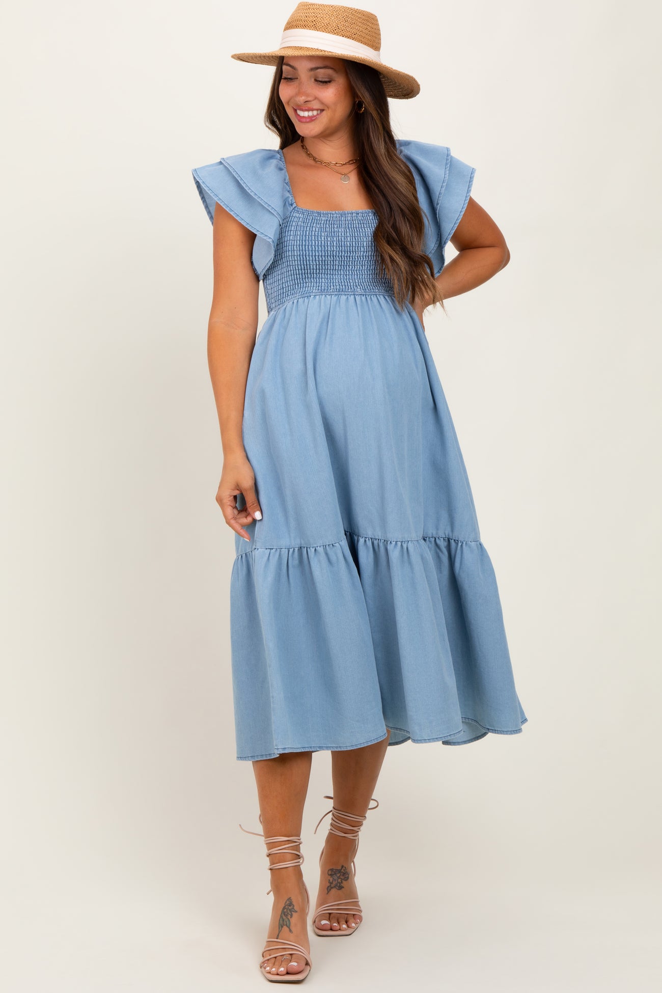 woman wearing a light blue maternity dress with ruffle sleeves and a smocked bodice, standing with a hat and sandals