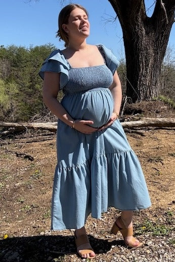 woman wearing a light blue maternity dress with flutter sleeves and a tiered skirt, holding her baby bump outdoors