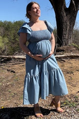 woman wearing a light blue maternity dress with flutter sleeves and a tiered skirt, holding her baby bump outdoors