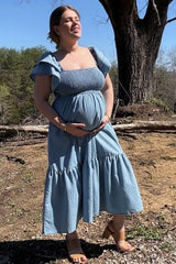 woman wearing a light blue maternity dress with ruffled sleeves and a tiered hem, holding her baby bump outdoors