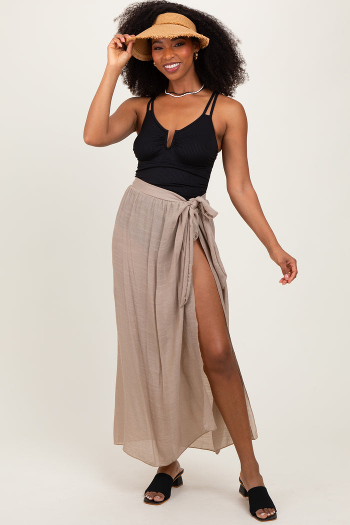 woman wearing a black ribbed swimsuit top and a tan wrap maxi skirt, accessorized with a straw hat and slip-on sandals