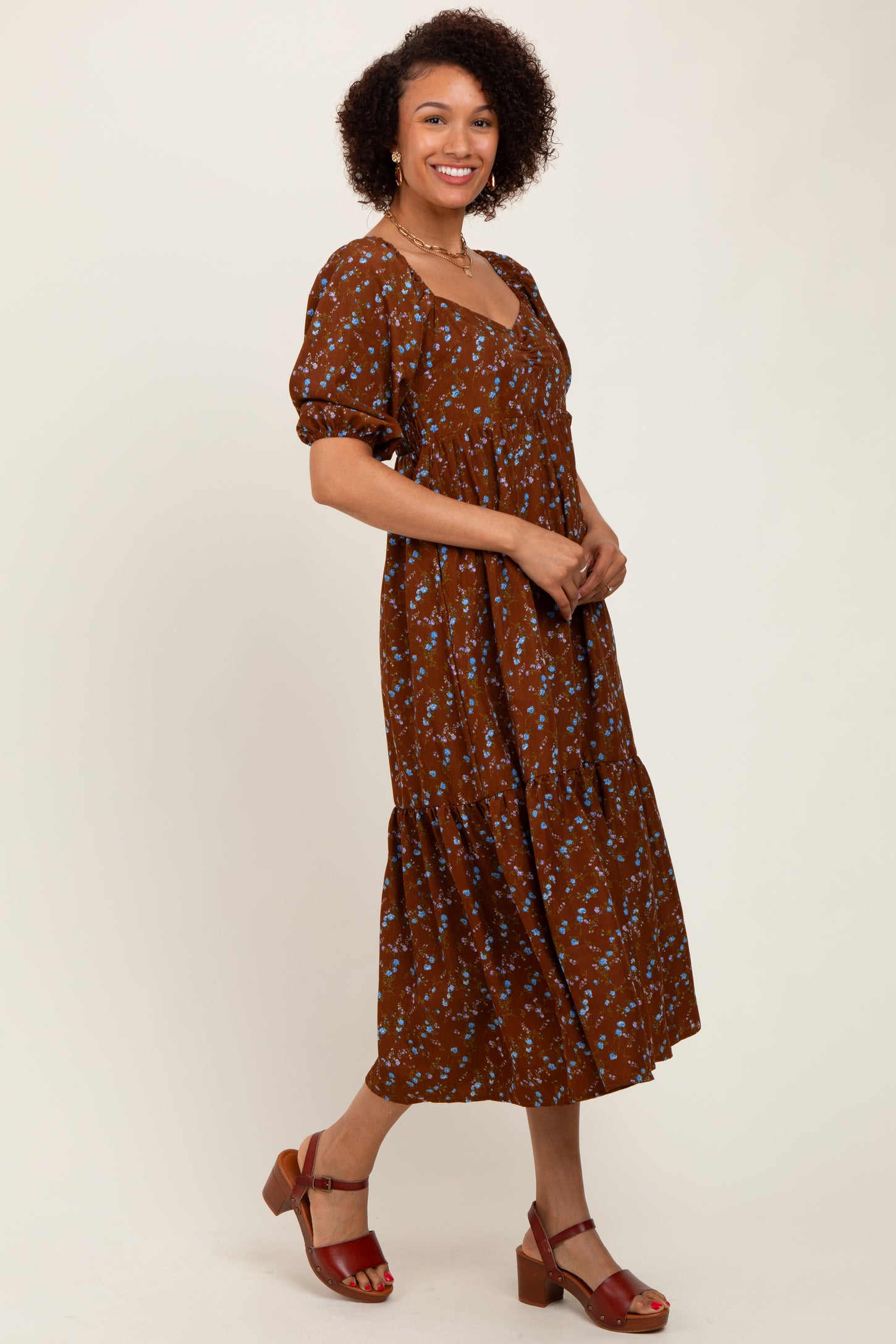 woman wearing a brown floral midi dress with puff sleeves and a tiered skirt, standing and smiling