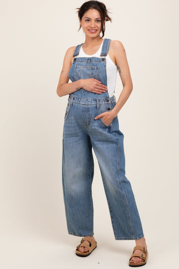 woman wearing a light blue maternity denim overall with wide legs and a white tank top, holding her baby bump