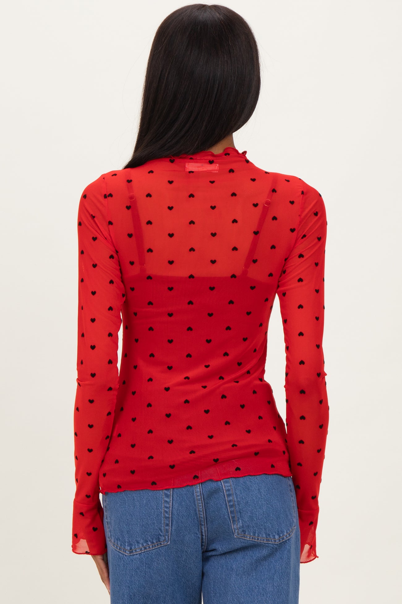 woman wearing a red sheer long sleeve top with black heart polka dots and a fitted back, paired with blue jeans