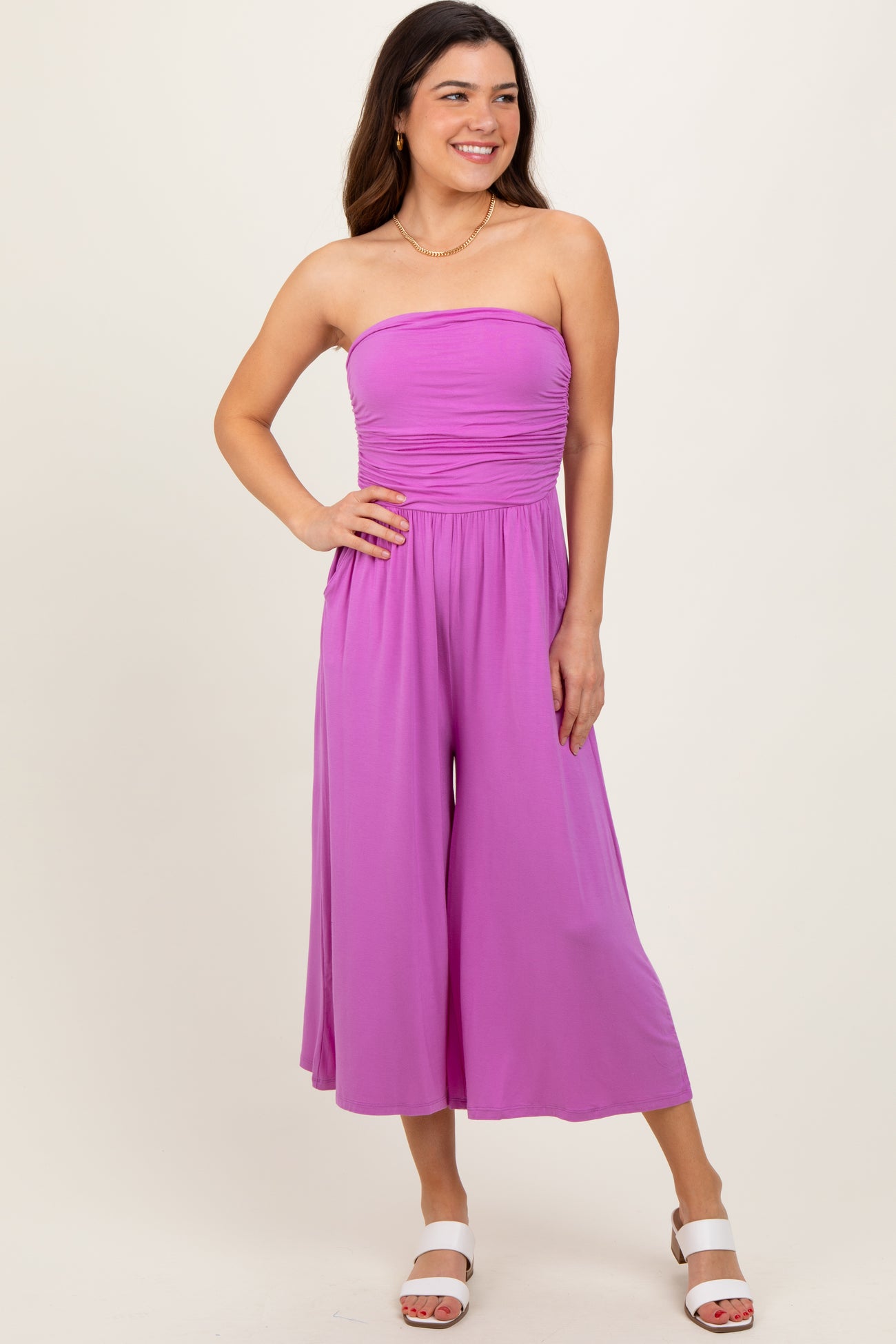 woman wearing a strapless purple jumpsuit with a ruched bodice and wide legs, standing and smiling
