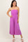 woman wearing a strapless purple jumpsuit with a ruched bodice and wide legs, standing and smiling