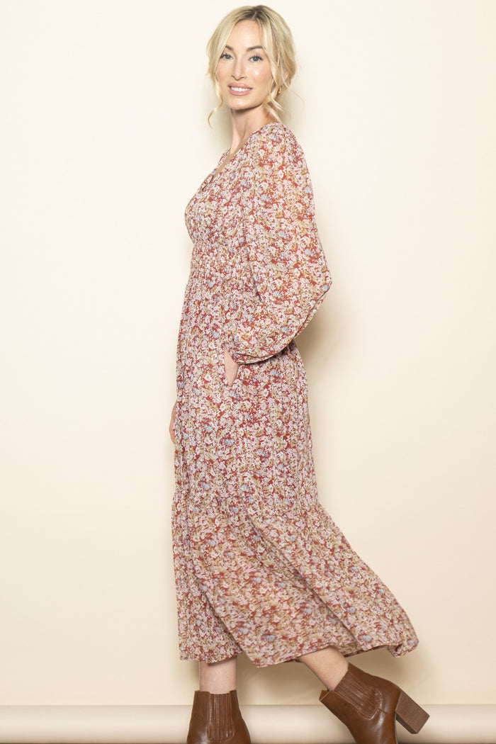 woman wearing a floral maxi dress with long sleeves and pockets, smiling against a neutral background