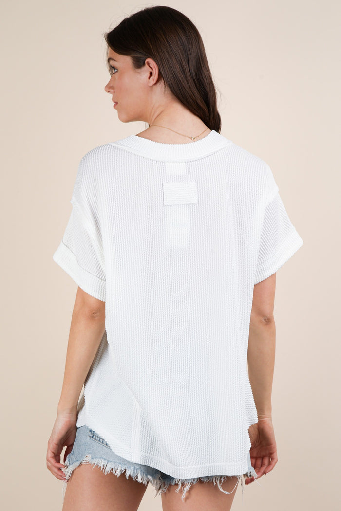 woman wearing a loose fit white knit top with rolled sleeves and back detailing, paired with denim shorts