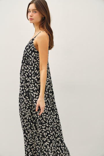 woman wearing a black maxi dress with white floral prints and spaghetti straps, posing sideways against a plain background