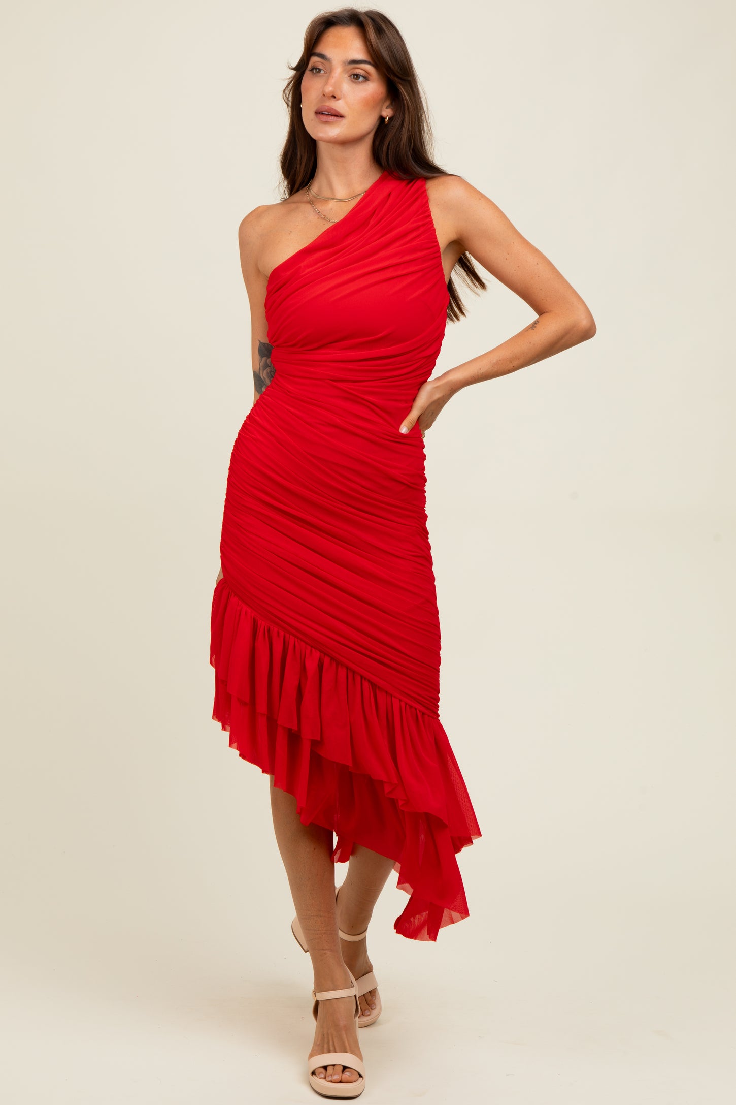 woman wearing a red one shoulder ruched dress with a ruffled hem line and standing with one hand on her hip