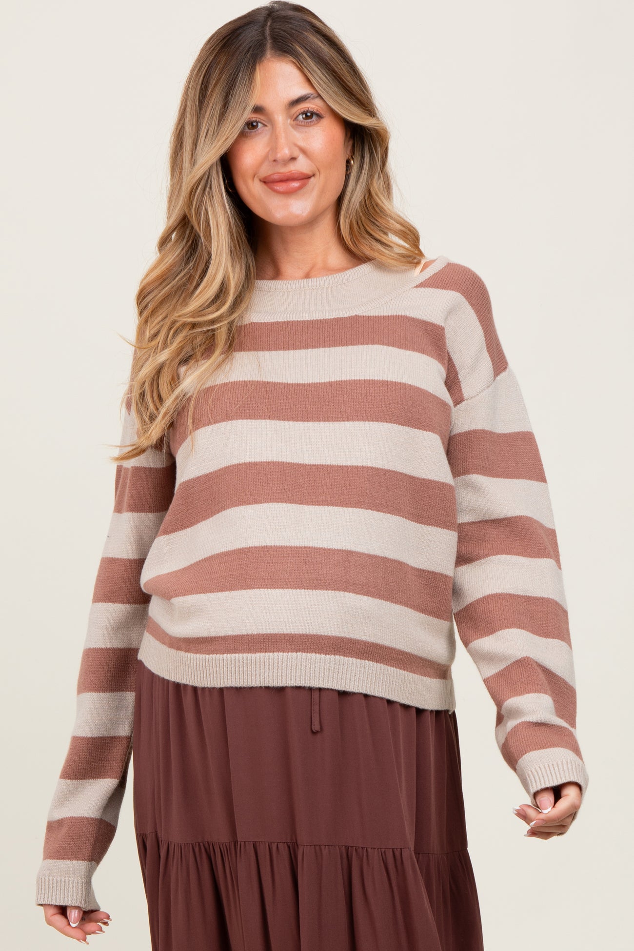 woman wearing a beige and brown striped maternity sweater over a brown tiered dress and smiling, revealing her baby bump