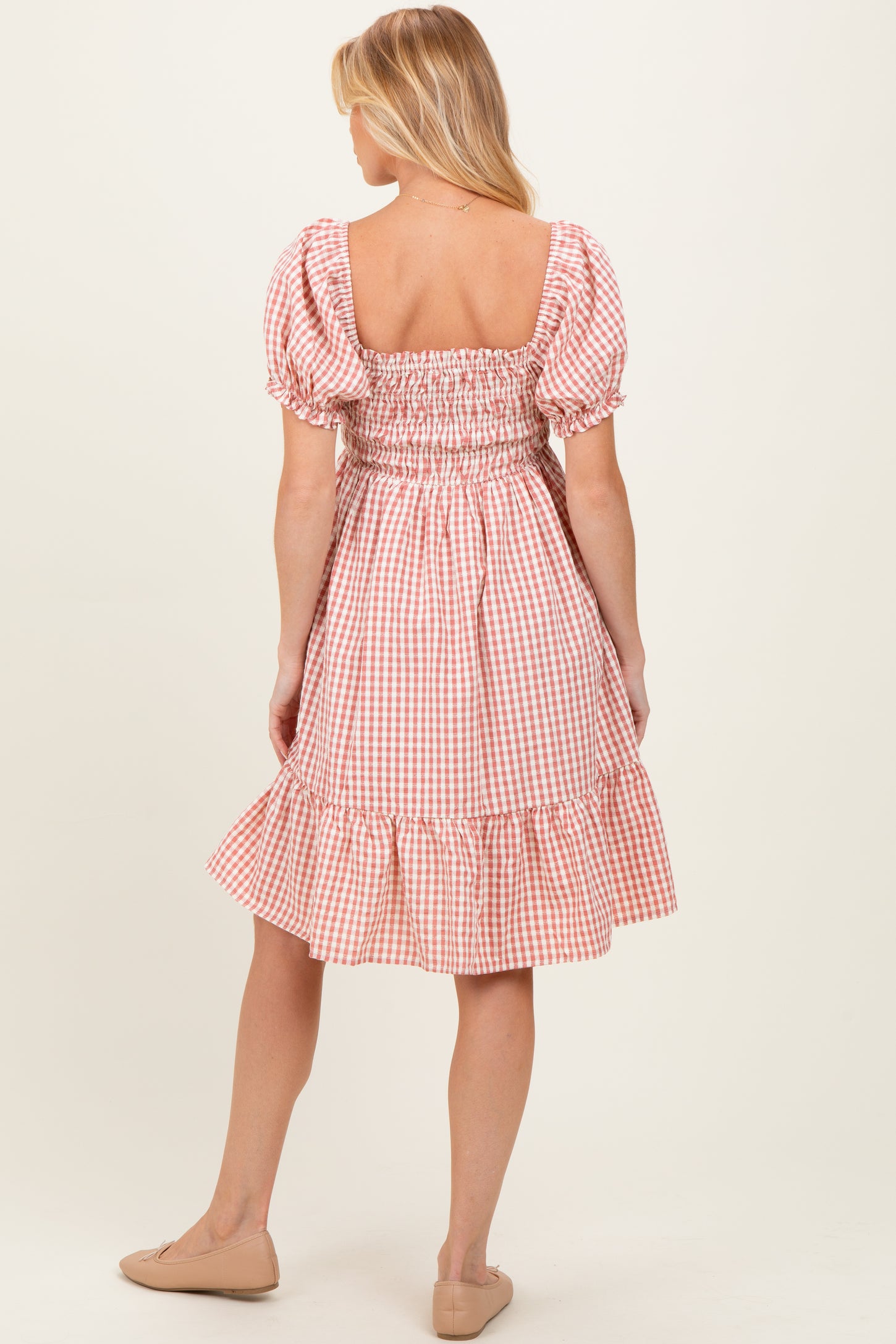 woman wearing a coral and white gingham maternity dress with puff sleeves and ruffle hem, showing her back
