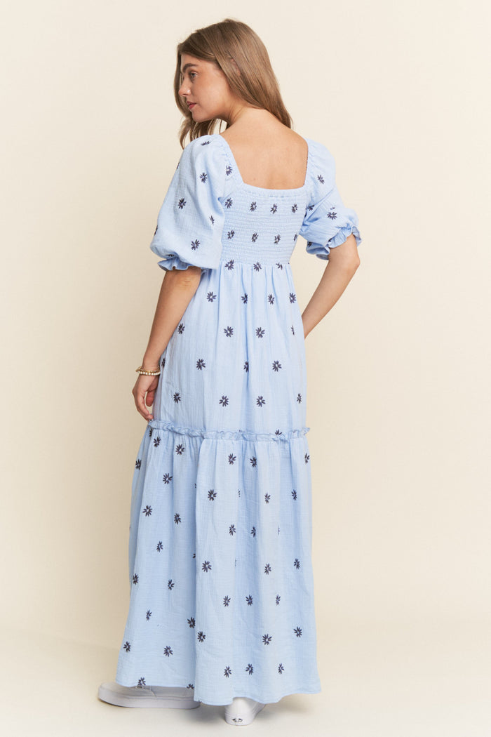 woman wearing a light blue floral embroidered maxi dress with puff sleeves and a smocked bodice, shown from the back