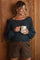 woman wearing a dark teal knit maternity sweater and brown checkered shorts, holding a white mug and smiling