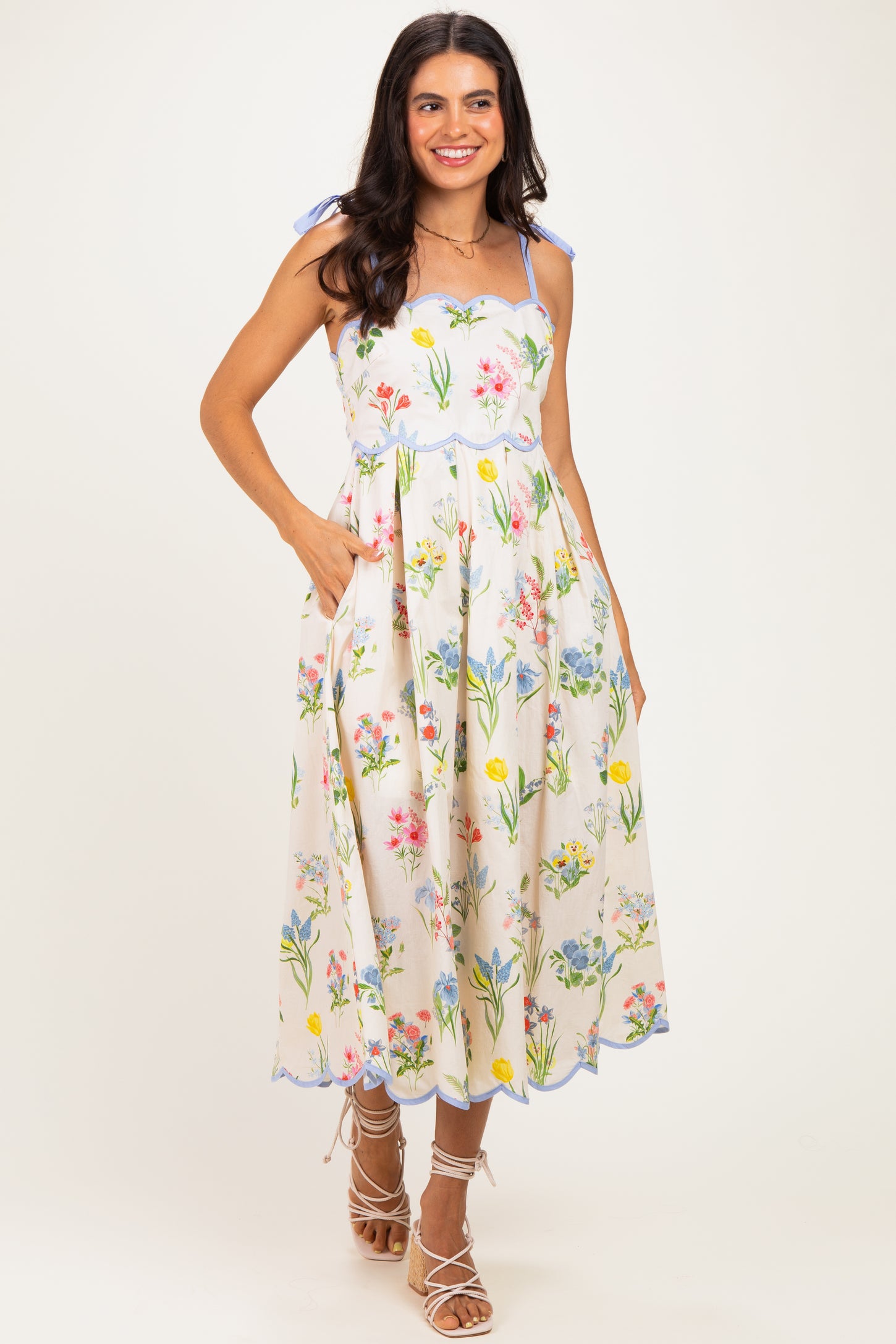 woman wearing a floral midi dress with spaghetti straps and pockets, featuring colorful flowers on a cream background